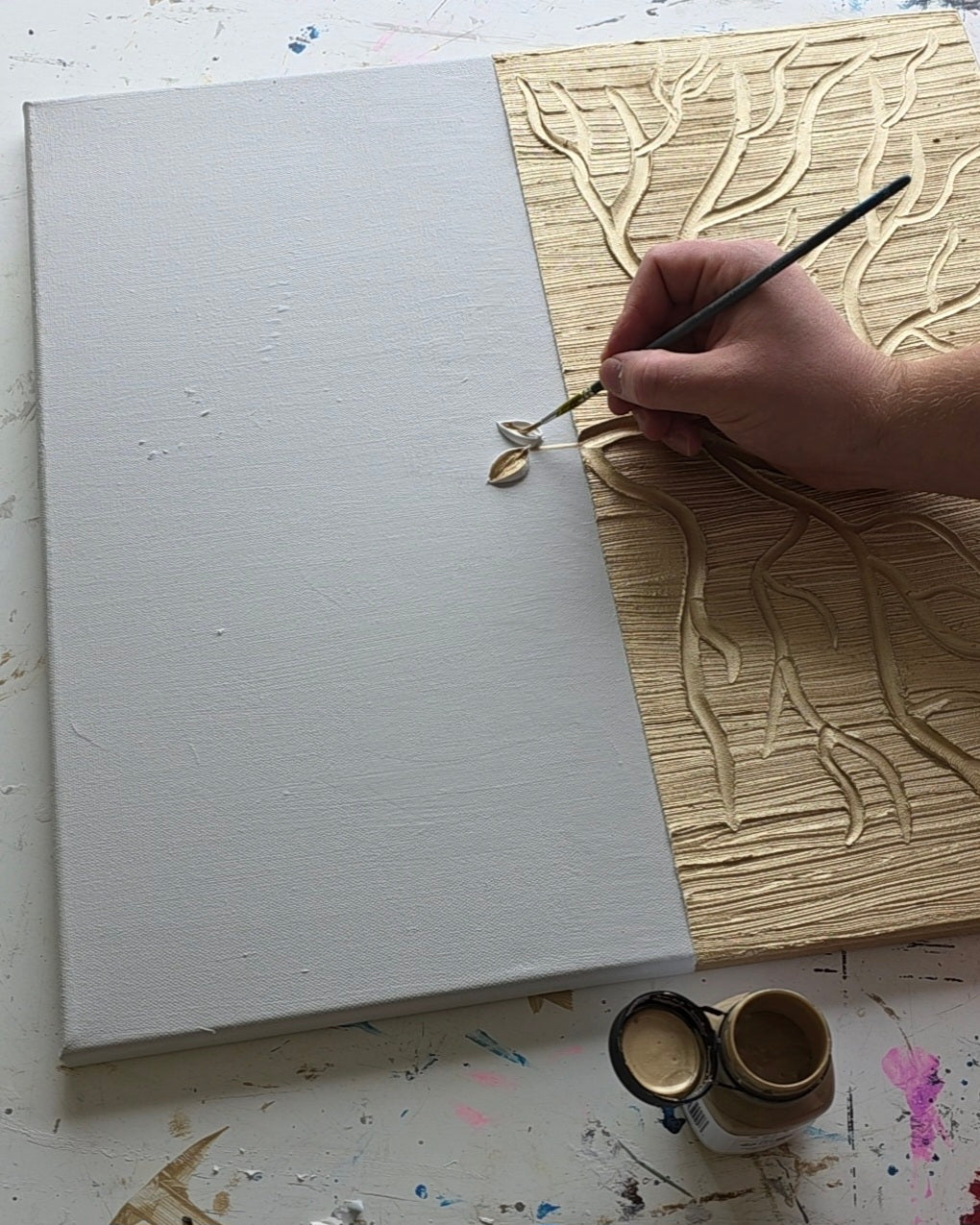 A hand holding a fine paintbrush applies gold paint to a canvas that is split vertically, with the left half blank and the right half showing textured gold tree branches. A small jar of gold paint sits on a paint-splattered white surface.